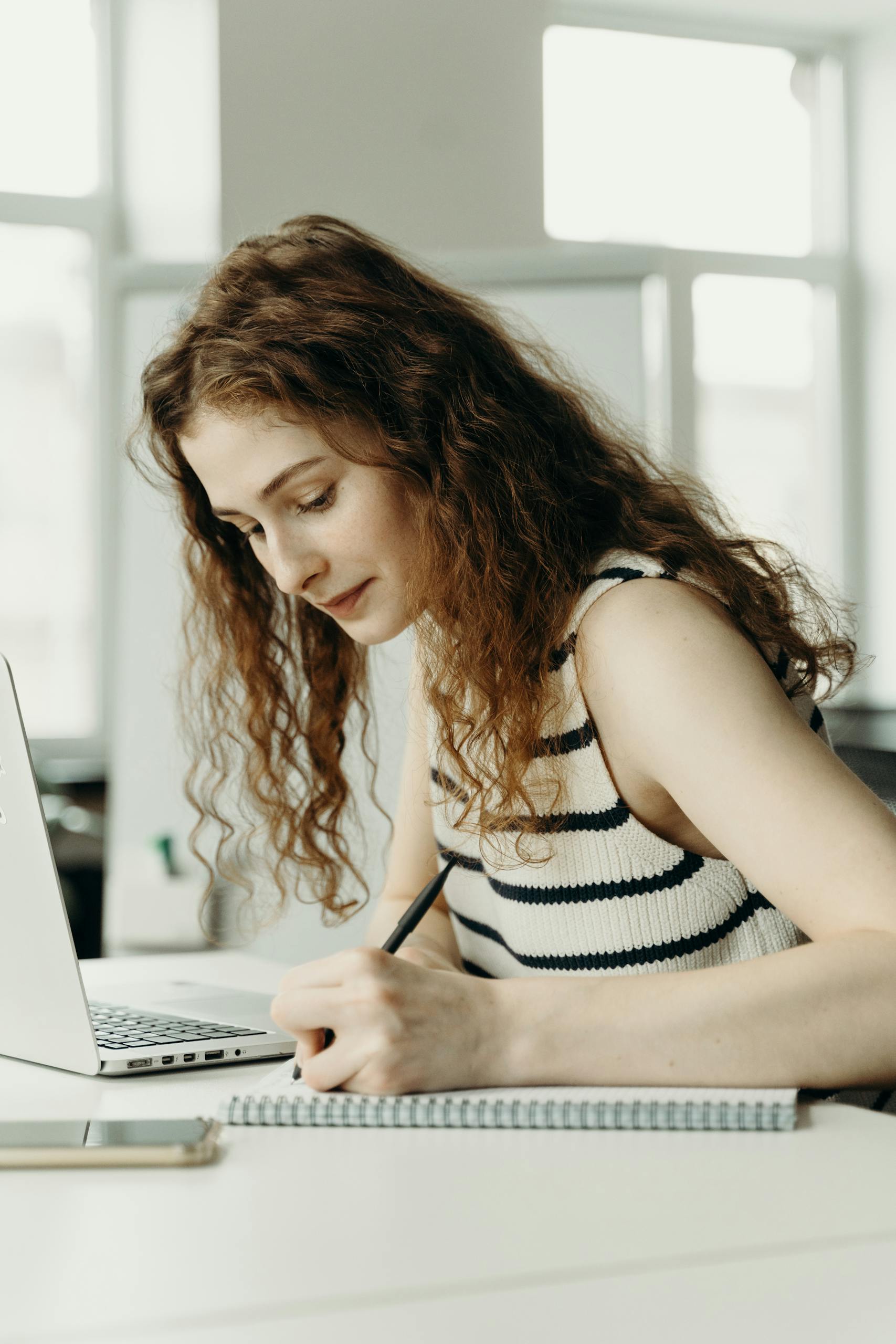 A professional woman writing notes at her laptop in an office setting.